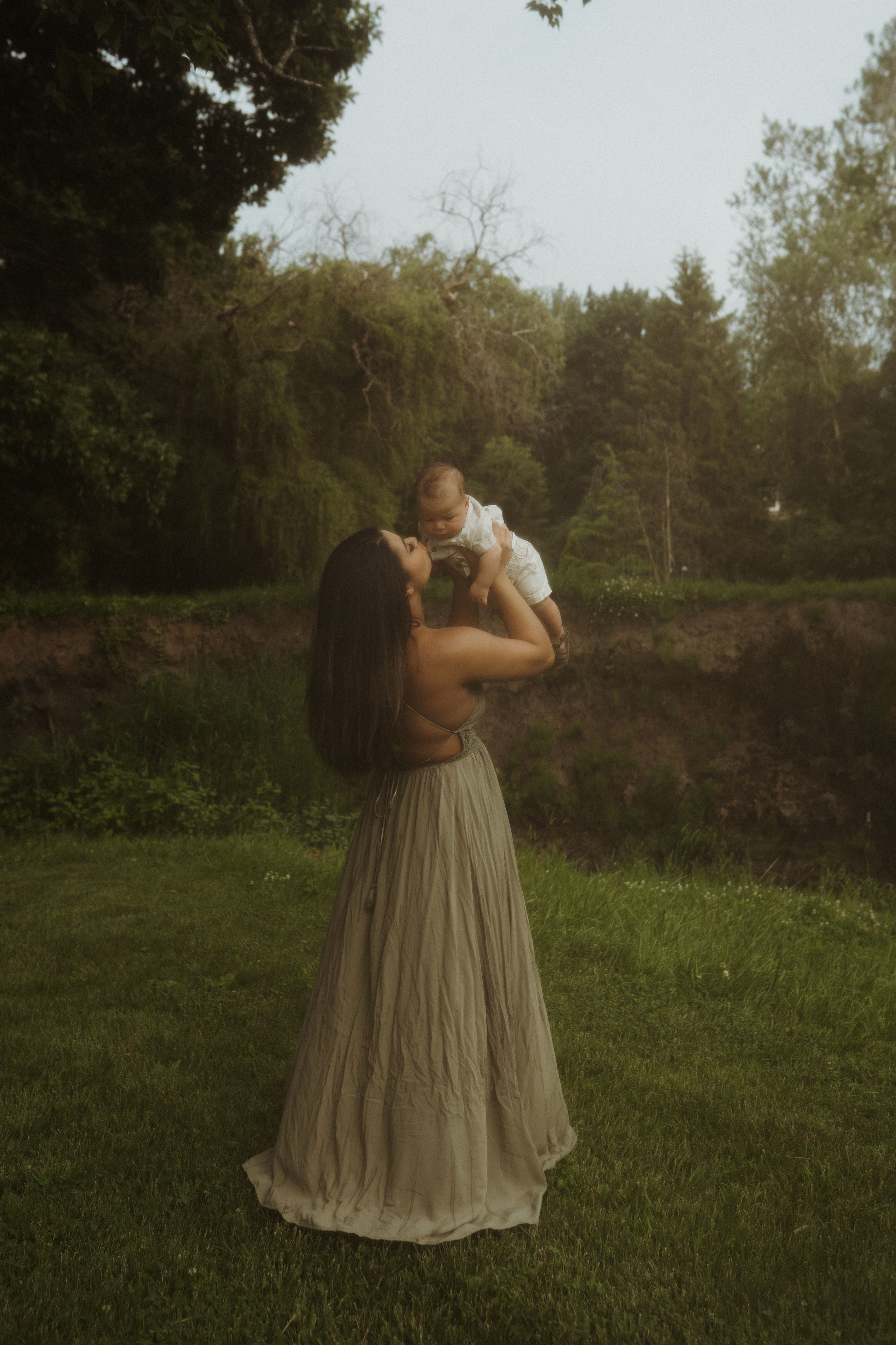 A tender moment captured in a garden setting as a figure in a flowing taupe dress lifts a small child against a wooded backdrop.