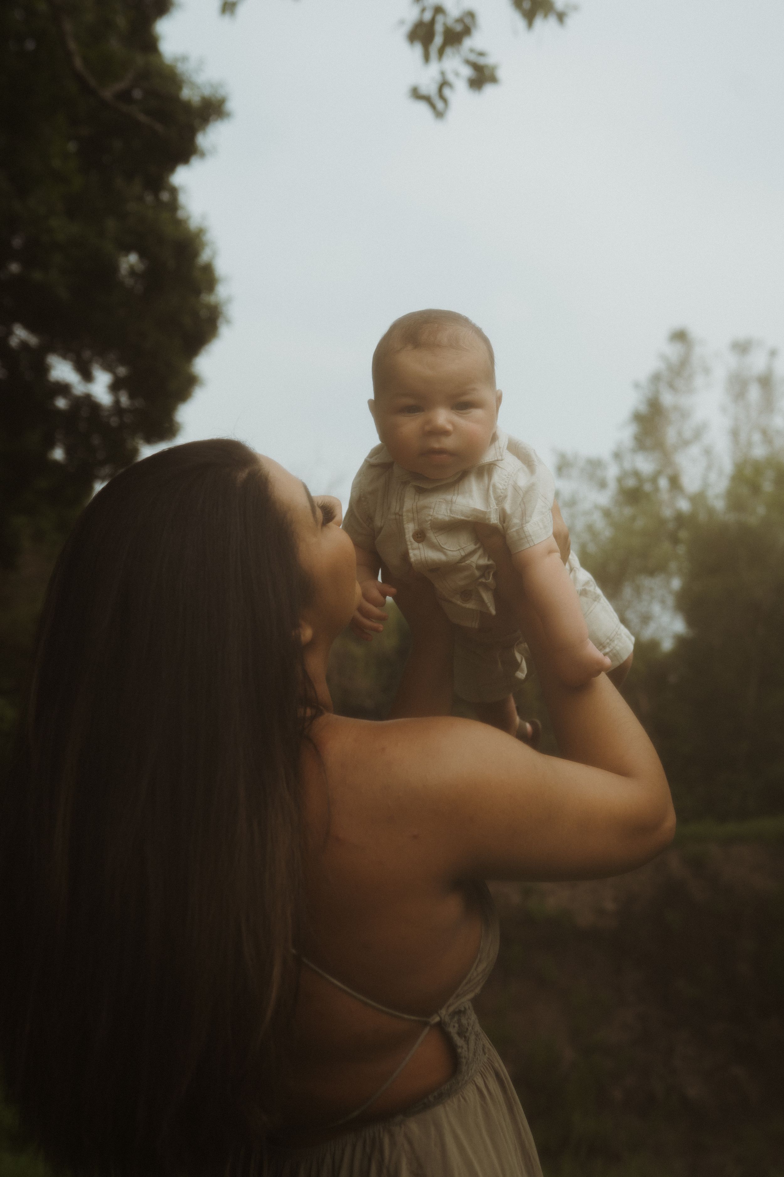 A person in a light-colored dress lifts a small infant up against a natural outdoor backdrop.