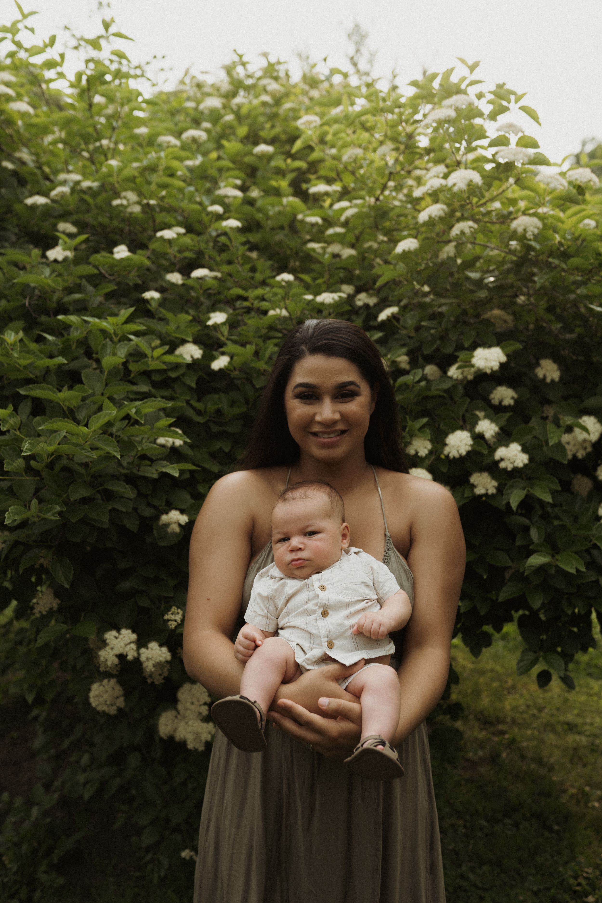 A spring portrait against blooming white elderflower bushes featuring a parent and infant in coordinated outfits.