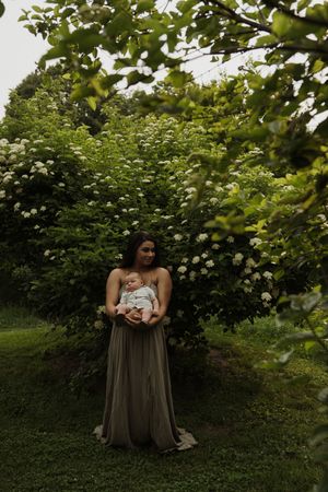 A serene garden portrait featuring flowing dress and baby against lush elderflower bushes.