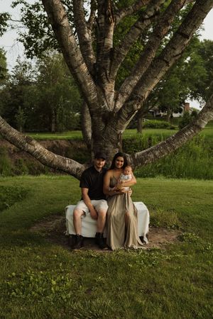 A couple sits together on a white blanket beneath a large birch tree in a grassy outdoor setting at dusk.