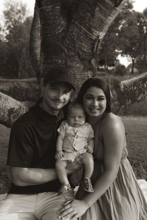 A family portrait in black and white showing a couple with their baby sitting under a large birch tree.