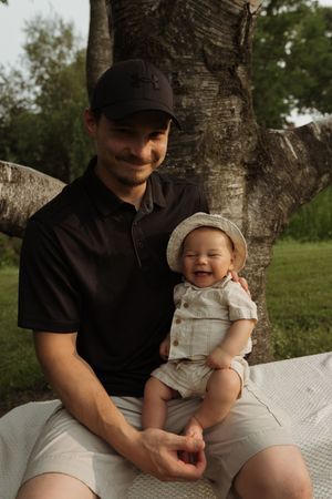 A cheerful moment between parent and baby sitting on a white blanket under a birch tree.