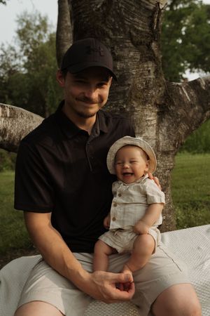 A parent and baby sit together outdoors on a blanket near a tree trunk in a natural setting.