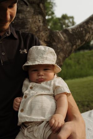 A close-up portrait of a baby wearing a bucket hat sitting with an adult under a tree.