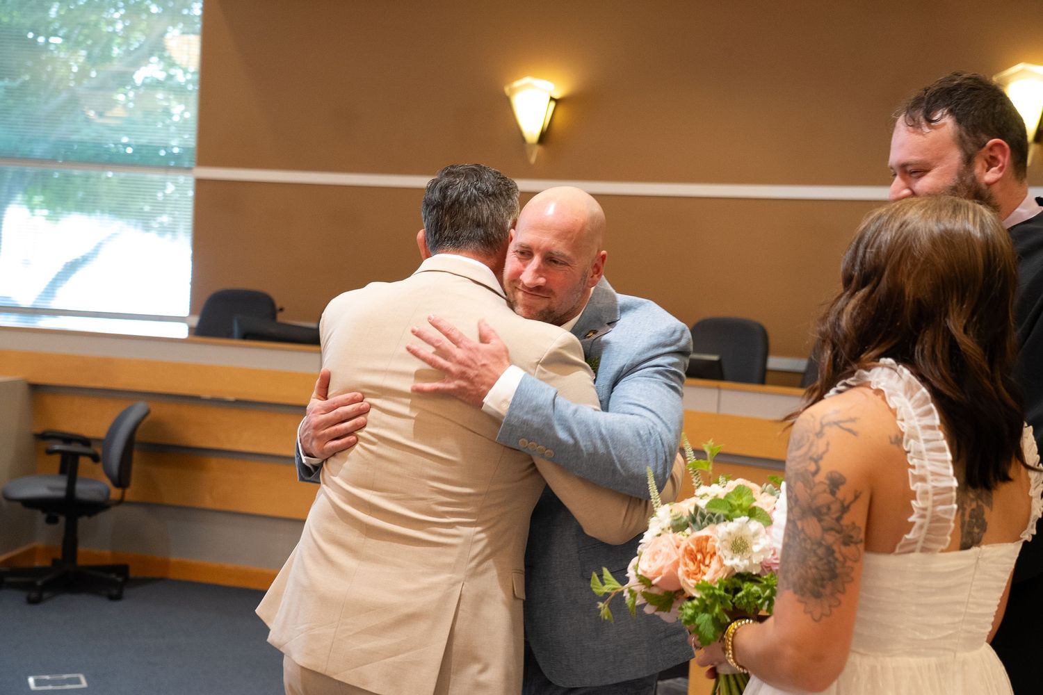 Two people embrace in an emotional moment at an indoor gathering while others look on with flowers nearby.