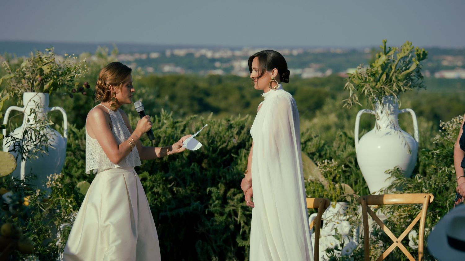 Two people in white attire share a special moment in an outdoor wedding ceremony with decorative urns and greenery.