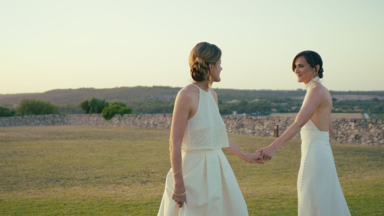 Two people in white wedding dresses hold hands while standing in a grassy field at sunset near stone walls.