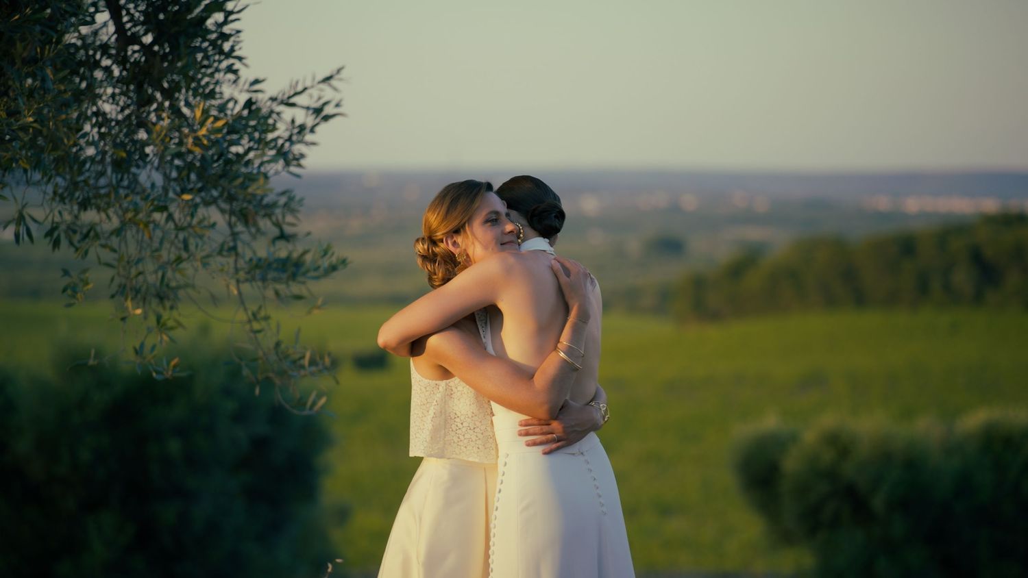 Two people in white dresses embrace on a hilltop overlooking a scenic landscape at dusk.