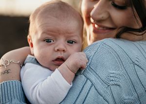A young infant in white clothing rests comfortably against someone wearing a blue knit sweater.