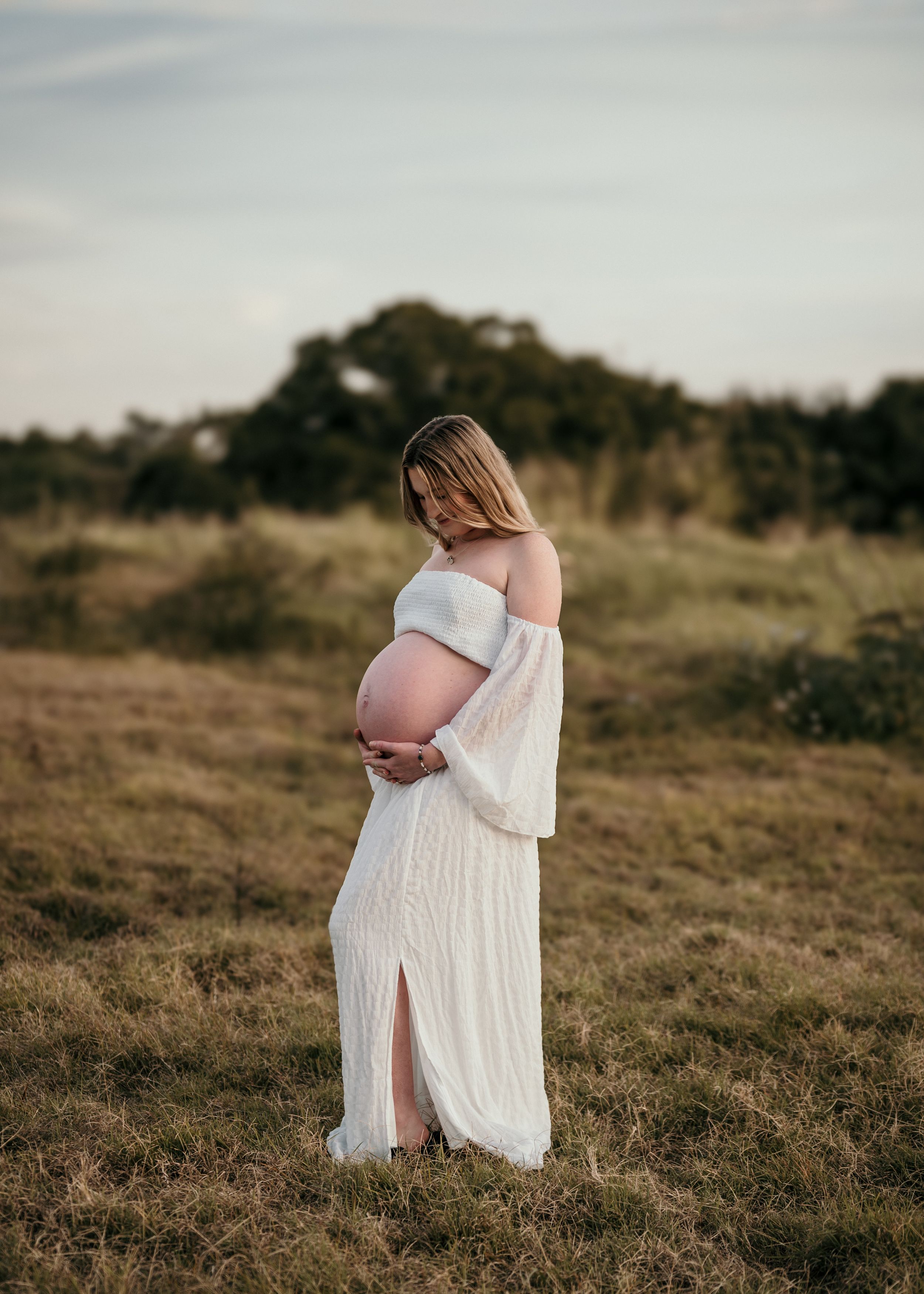 A pregnant figure stands in a flowing white dress against a natural outdoor landscape at golden hour.