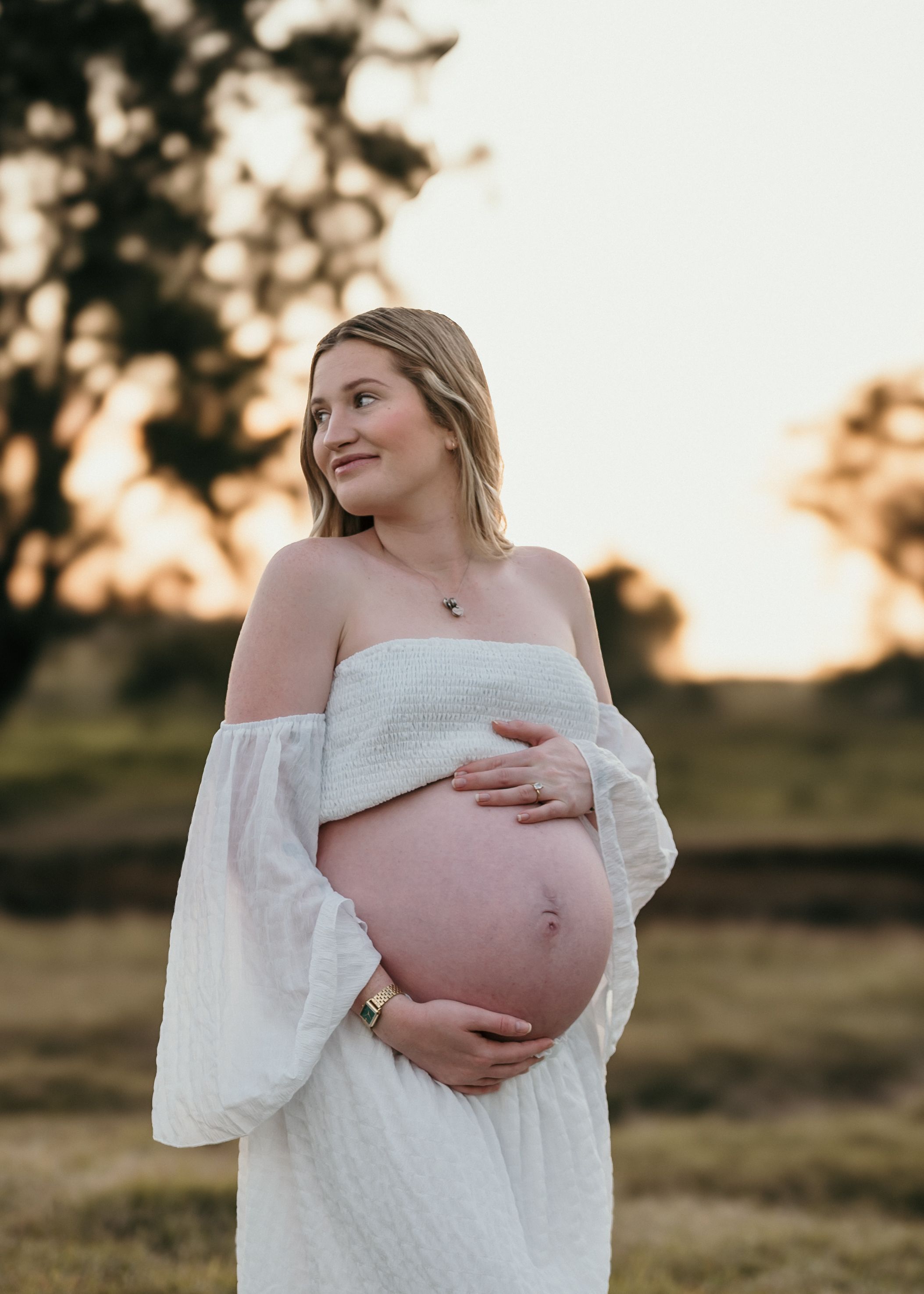 An expecting mother in a white off-shoulder dress cradles her baby bump during golden hour in a peaceful outdoor setting.