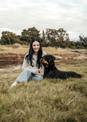 A person in casual clothing sits on grassy ground beside a black Labrador retriever near a pond on an autumn day.