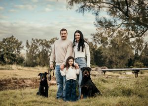 Two black dogs pose outdoors in a grassy field with three people on a sunny afternoon.