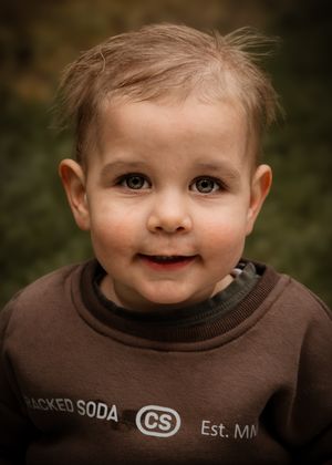 Young child wearing a brown sweatshirt smiles warmly at the camera in an outdoor portrait setting with soft lighting.