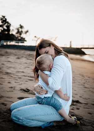 Tender embrace between parent and child sitting on a sandy beach at sunset with sea and palm trees in background.