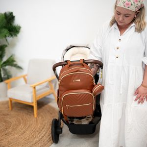 A brown leather baby stroller diaper bag is showcased next to a wooden armchair on a round jute rug.