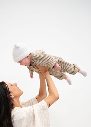 A woman in a white top lifts a baby wearing a striped outfit and white hat against a light background.