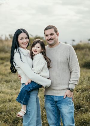 A family in matching cream sweaters and jeans embraces while standing in a grassy meadow during an outdoor photo session.