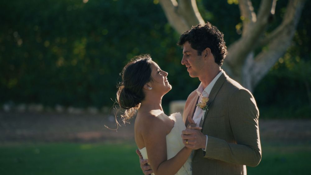 Romantic couple in formal attire dancing together during golden hour outdoors with trees in background.