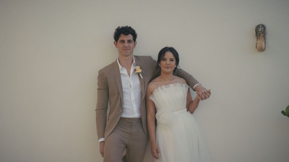 A couple poses against a white wall, with one person wearing a tan suit jacket and the other in a white wedding dress.