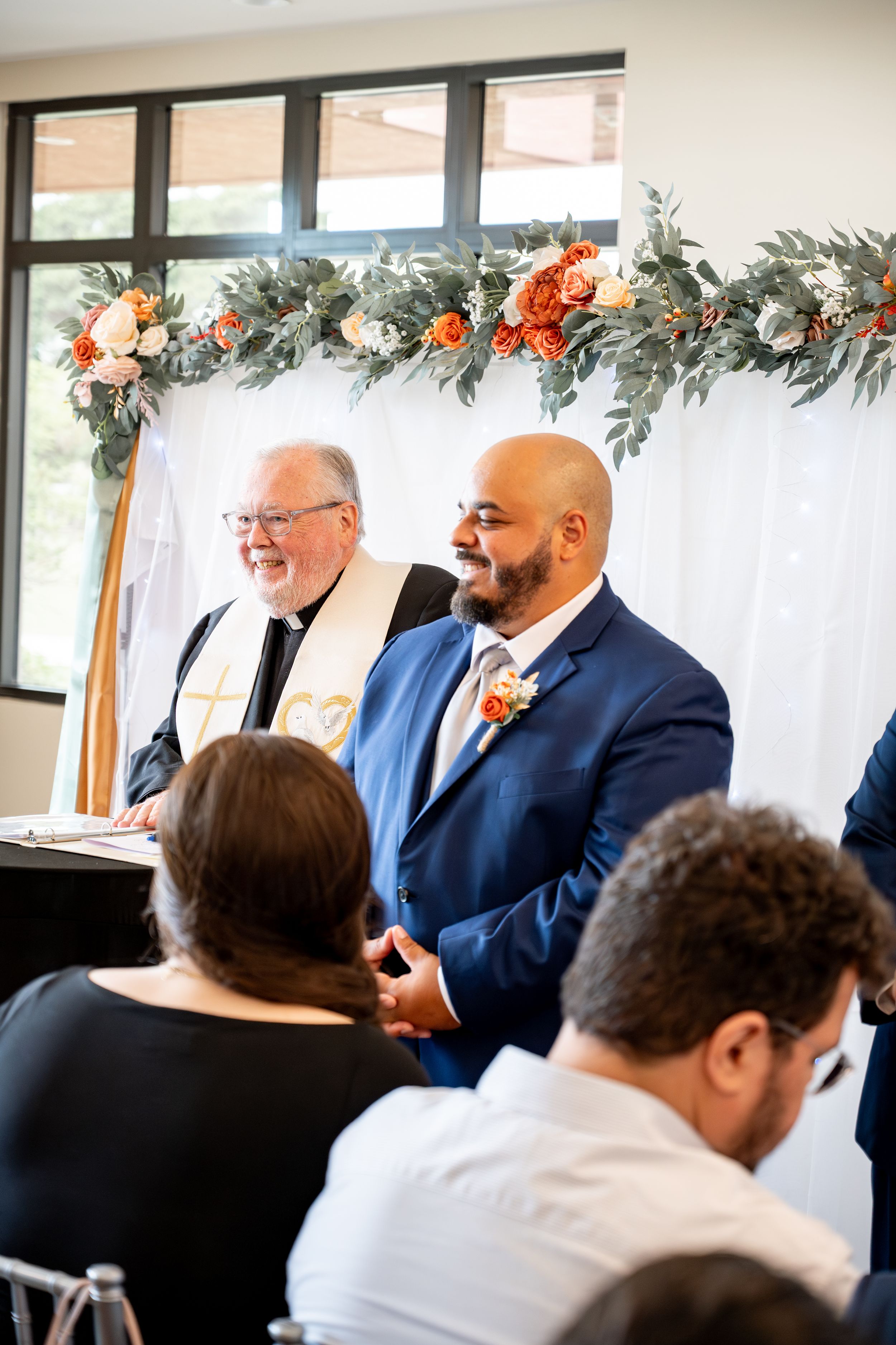 Wedding ceremony setup with floral decorations and white backdrop in an indoor venue.