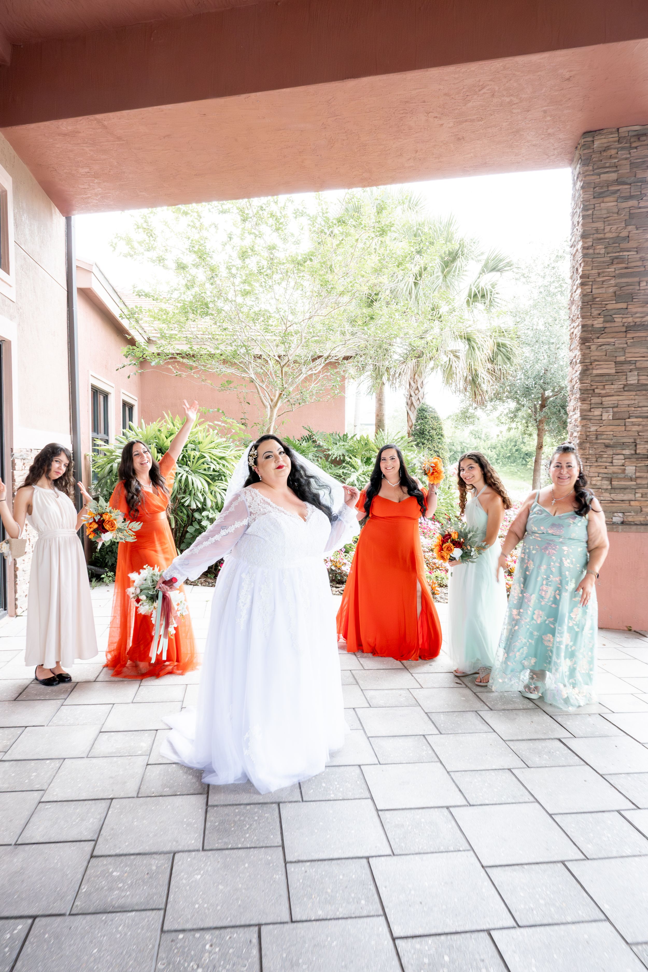 Bridal party celebrates on stone patio with bride in white and bridesmaids in orange and mint dresses.