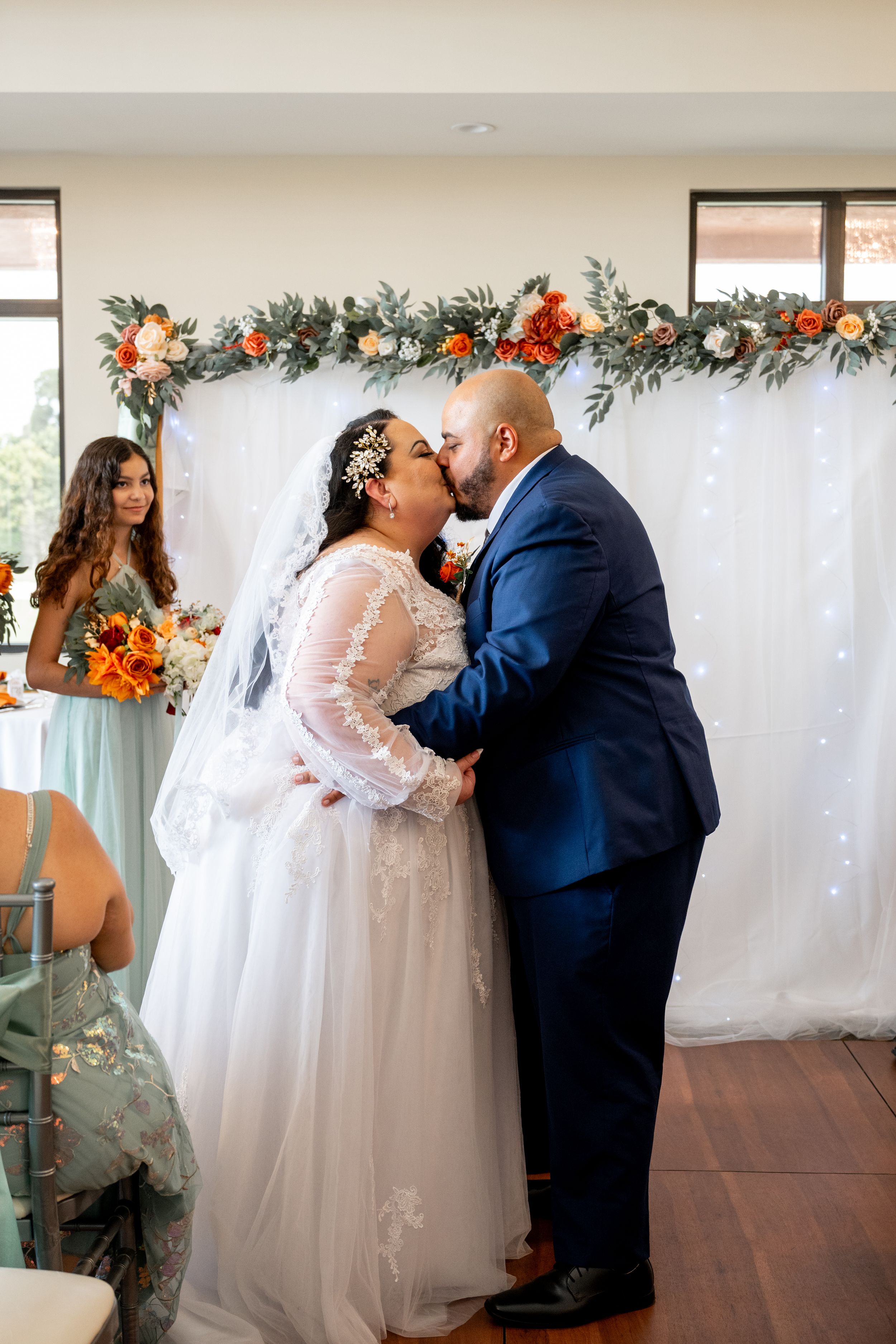 Wedding ceremony in an indoor venue decorated with floral garlands and white backdrop.