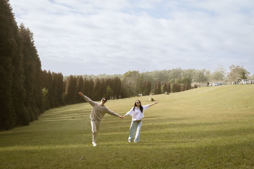 Two people holding hands and leaping joyfully across a green hillside on a cloudy day.