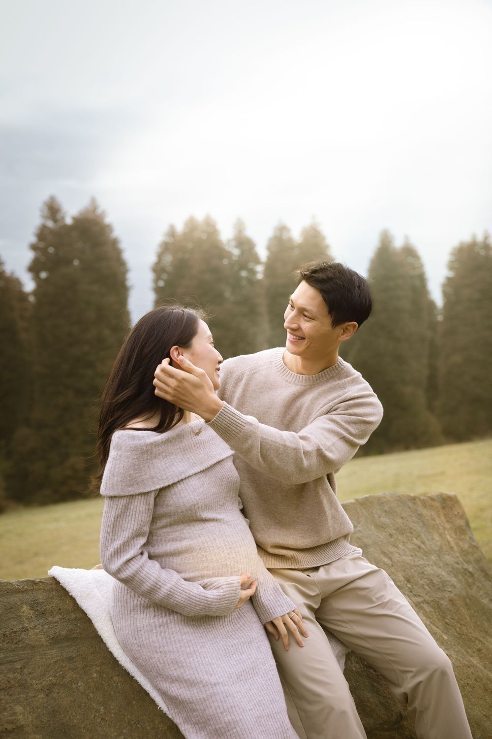 A couple in cozy sweaters share a playful moment together on a grassy hillside with evergreen trees in the background.