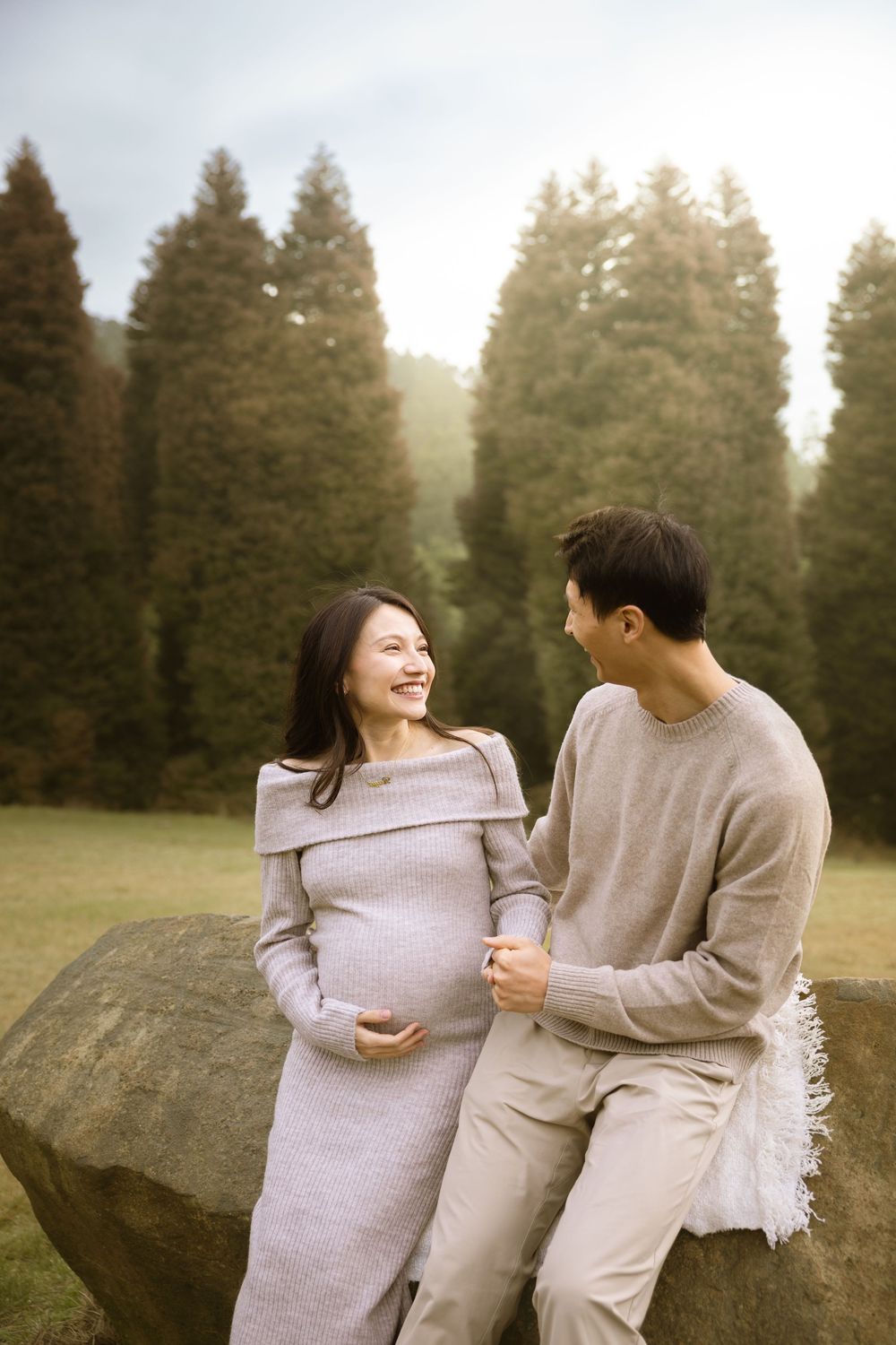 Expecting couple shares a joyful moment while sitting on a large rock in a forest setting.