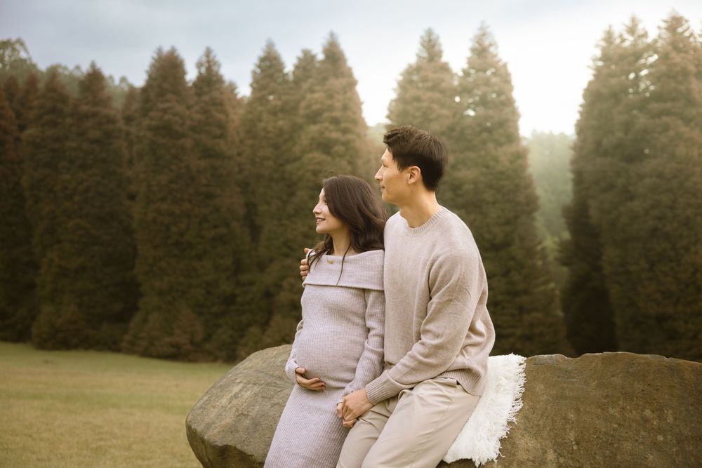 A couple stands together in a scenic forest setting during sunset, creating a romantic maternity photoshoot atmosphere.
