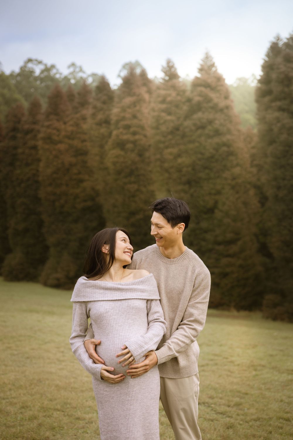 A couple in warm sweaters stands together in a grassy field with evergreen trees in the background at sunset.