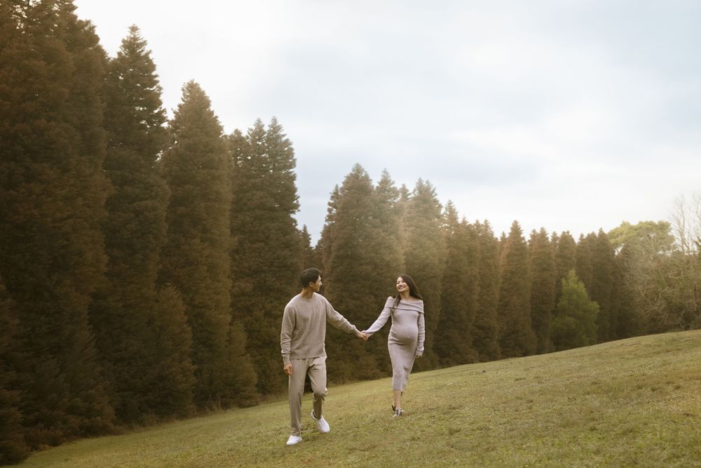 A couple strolls hand in hand across a grassy hillside with tall evergreen trees in the background at sunset.