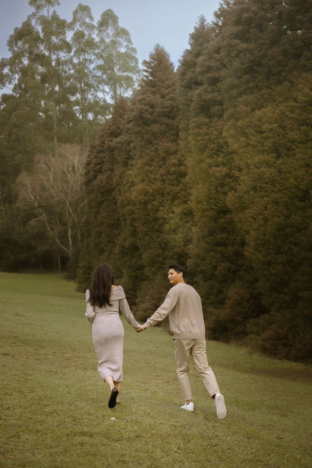 A couple holding hands runs across a grassy field surrounded by tall eucalyptus and pine trees.