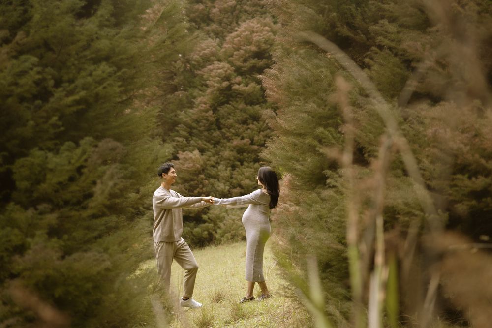 A couple holds hands and spins playfully on a grassy hillside surrounded by lush greenery.