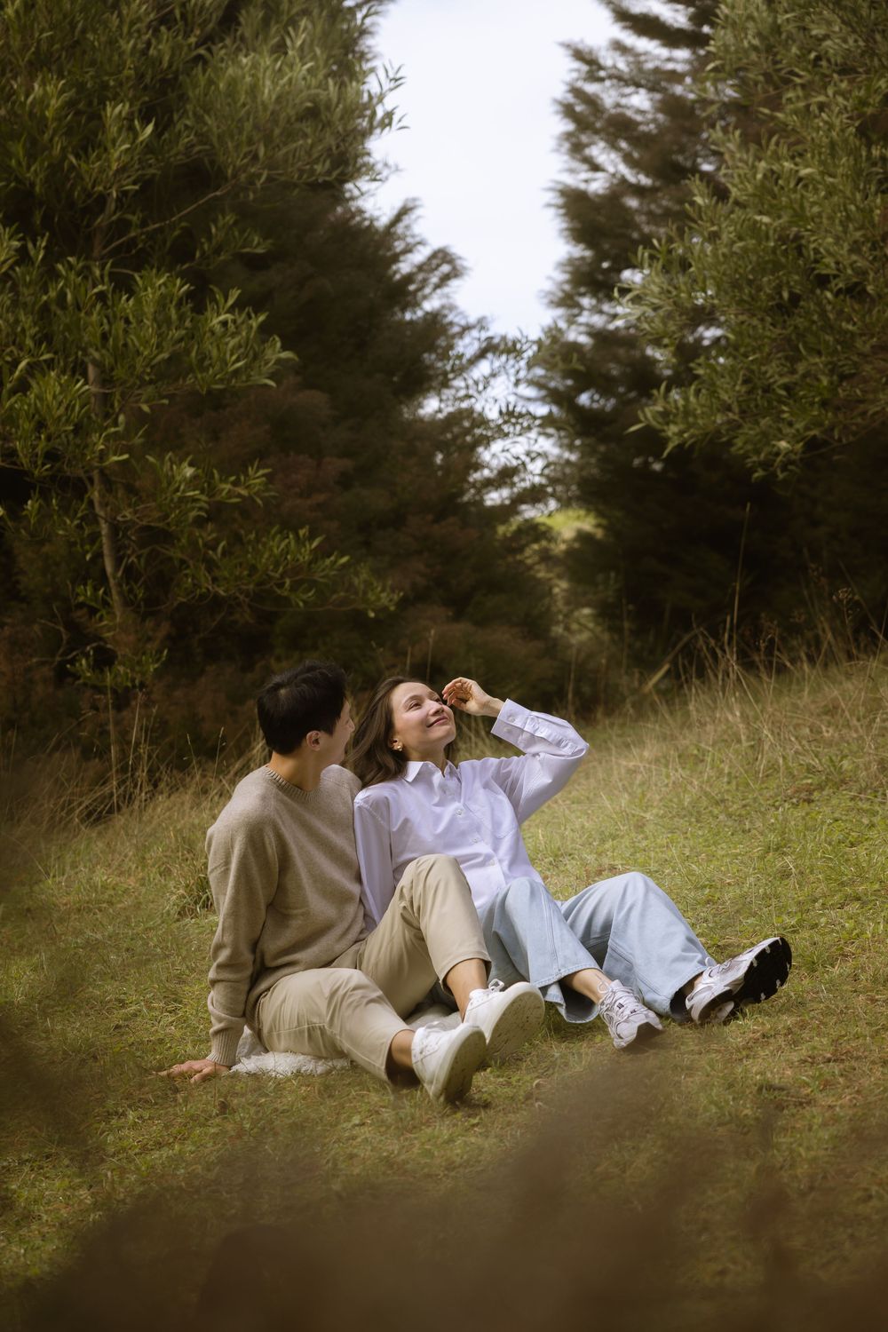 A couple relaxes on grassy ground surrounded by evergreen trees on a peaceful afternoon.