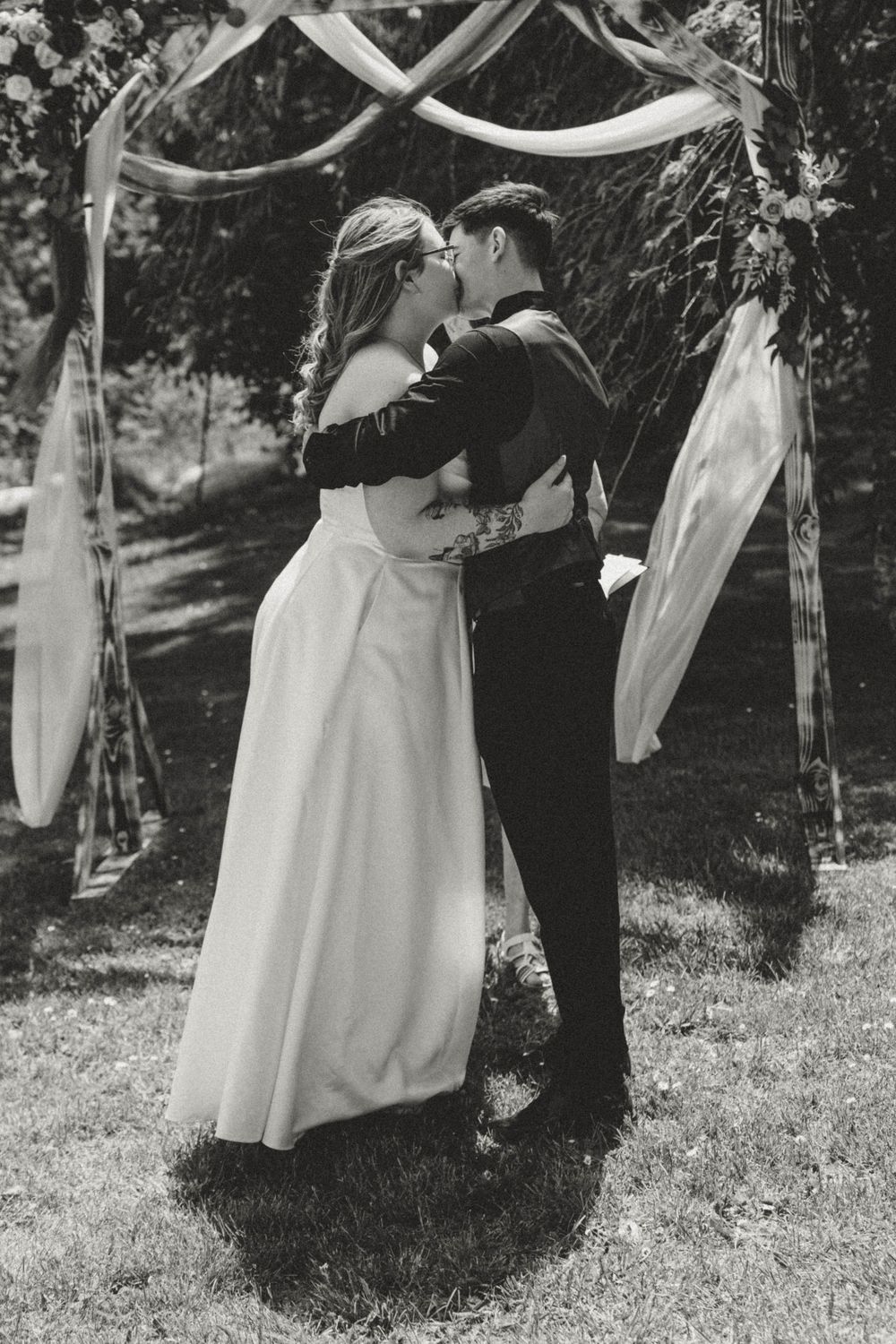 Black and white photo of newlyweds embracing under a floral wedding arch outdoors.