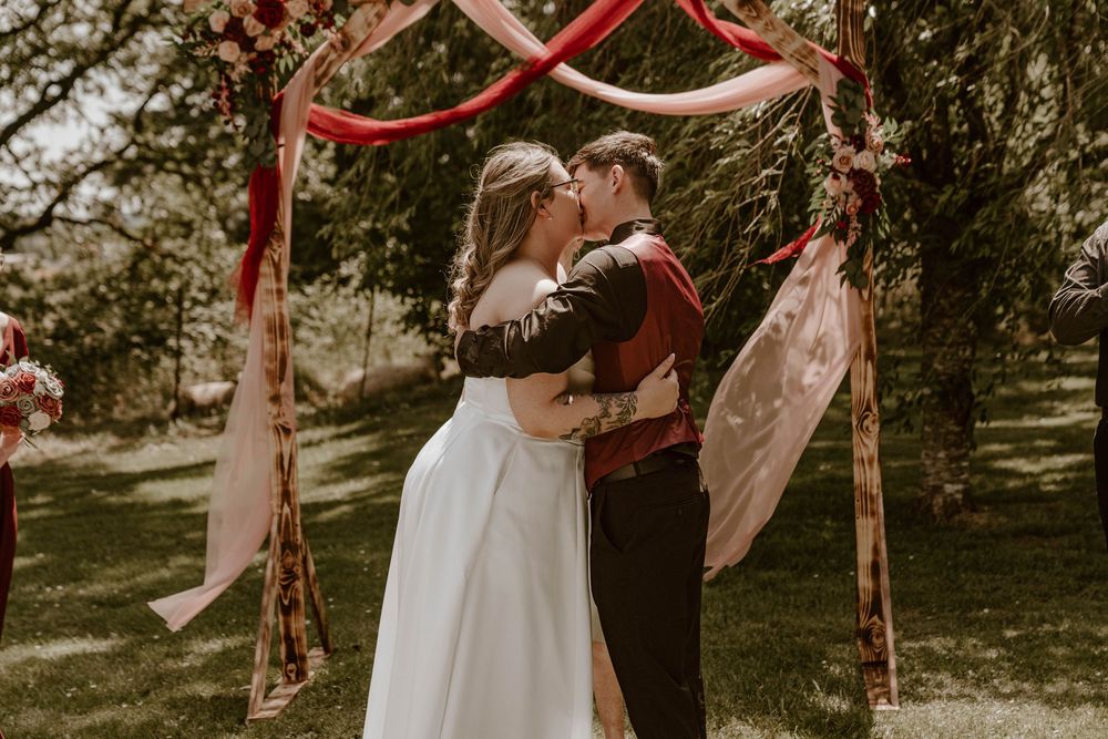 Bride and groom sharing their first kiss under a red draped floral wedding arch.