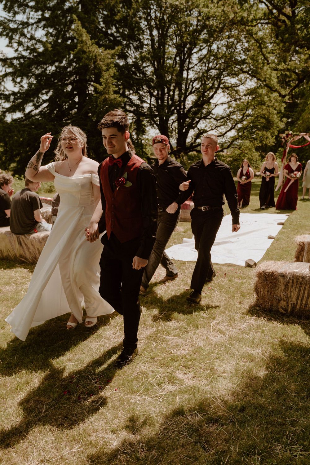 Wedding party walking down grass aisle with hay bales at outdoor rustic ceremony on sunny day.