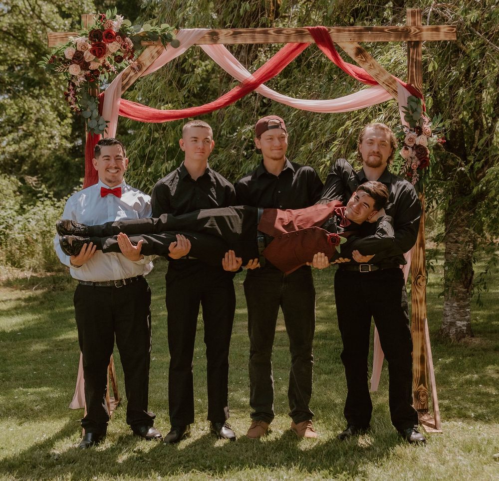 Five groomsmen in black suits posing with toy guns under floral wedding arch with red decorations.