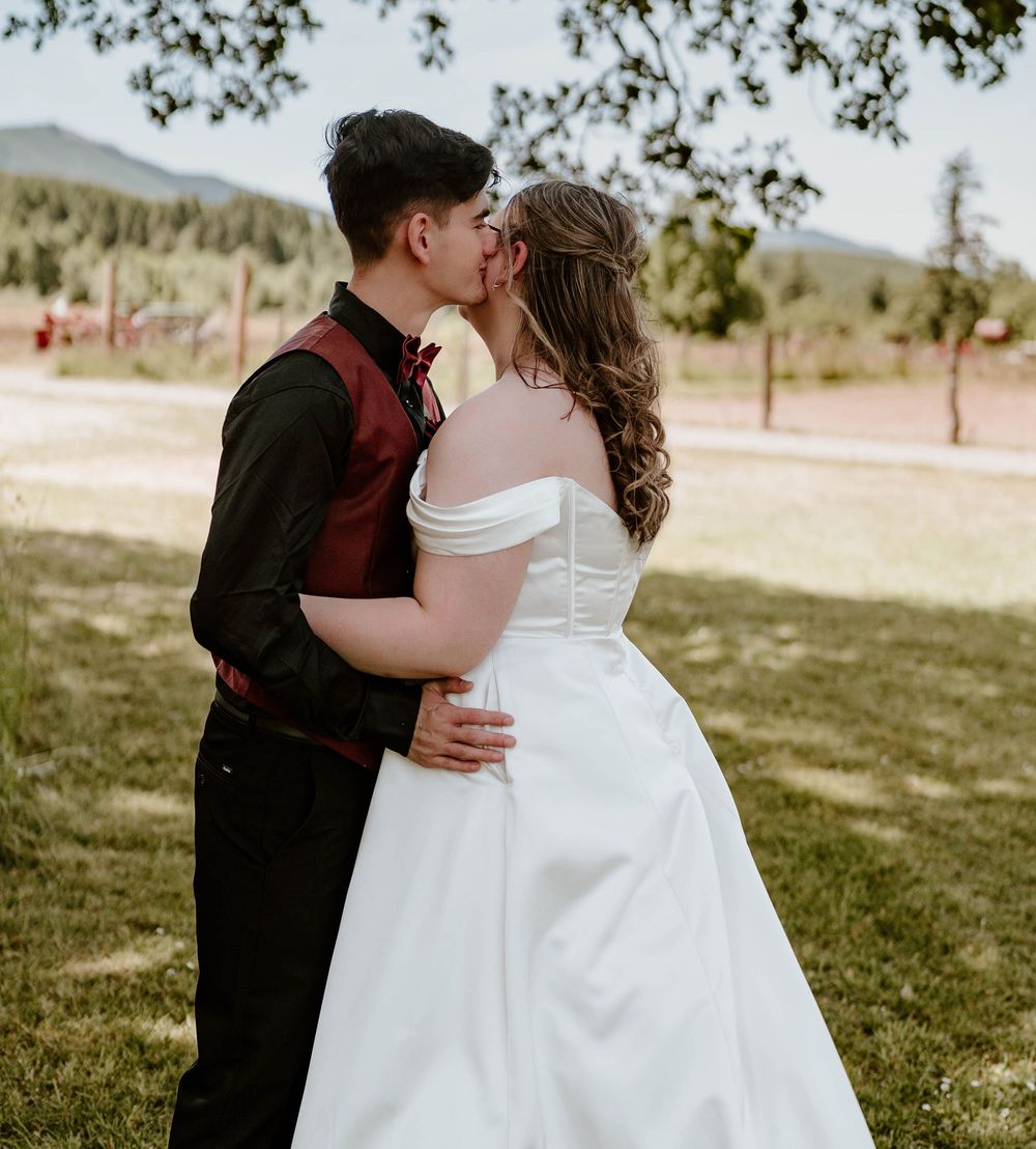 Bride and groom sharing first kiss in white off-shoulder gown and burgundy vest at outdoor wedding.