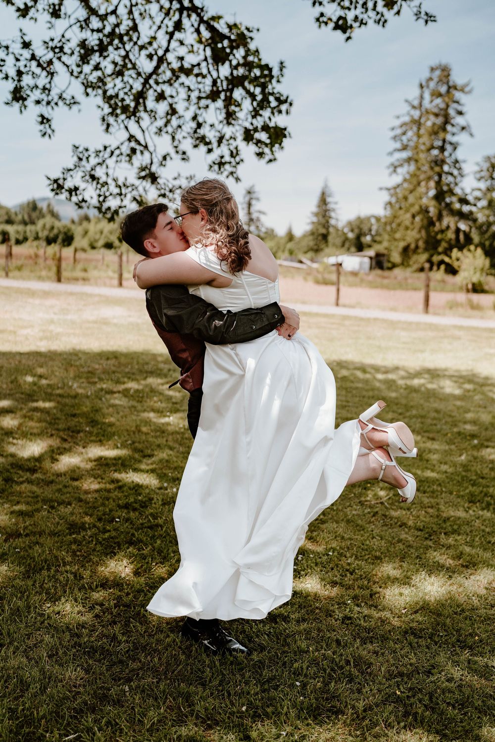 Groom lifting bride in white wedding gown for romantic kiss under tree at outdoor garden wedding.