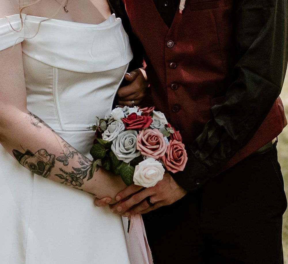 Close-up of bride and groom holding pink and red rose bouquet, bride showing floral arm tattoo.