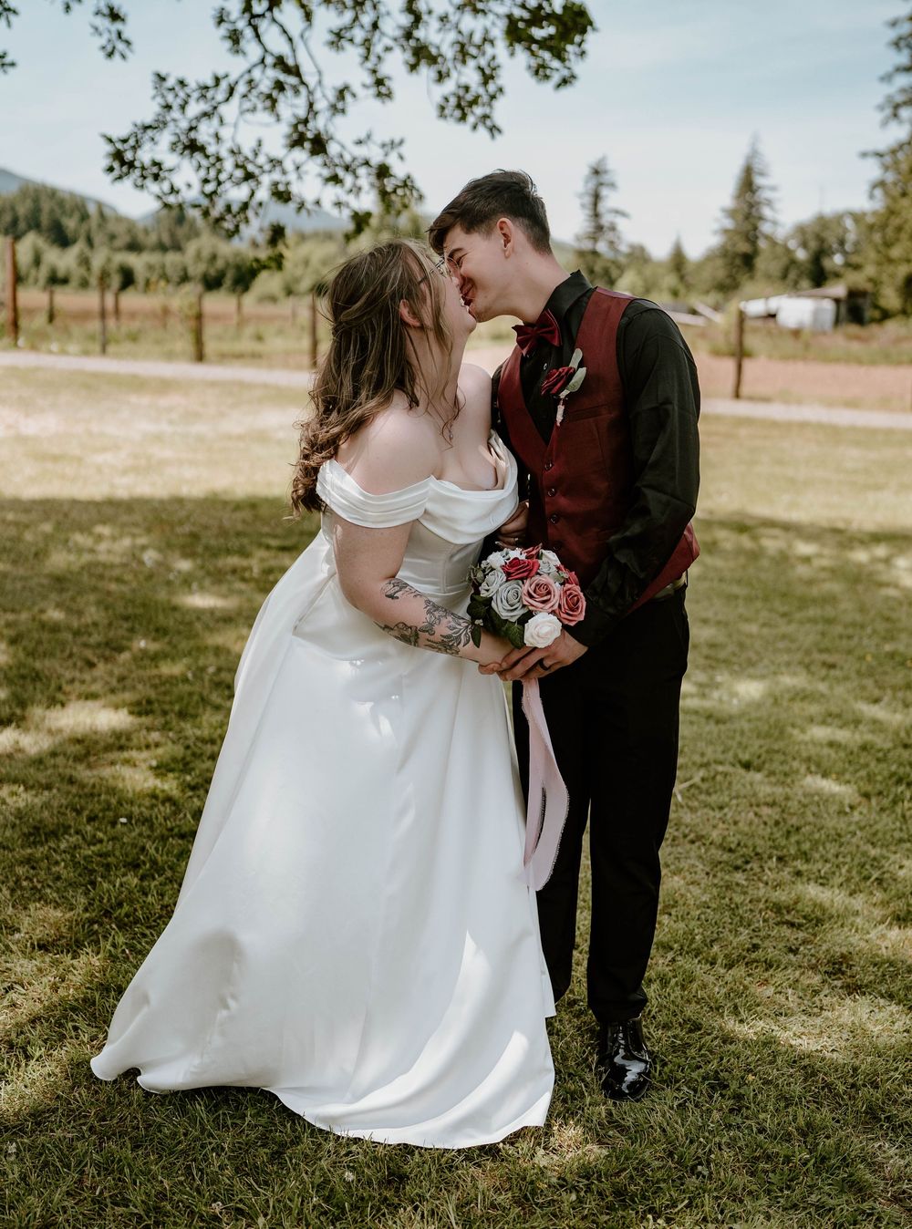 Bride and groom kissing in garden, bride in off-shoulder white gown holding pink and red bouquet.