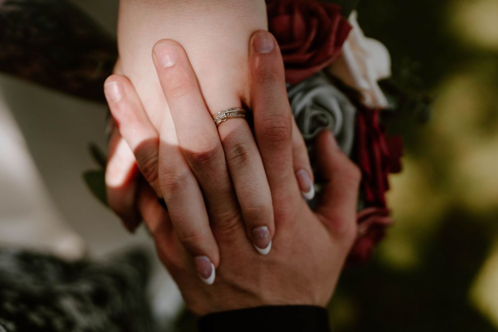 Close-up of newlyweds hands with wedding rings resting on red and white floral bouquet.
