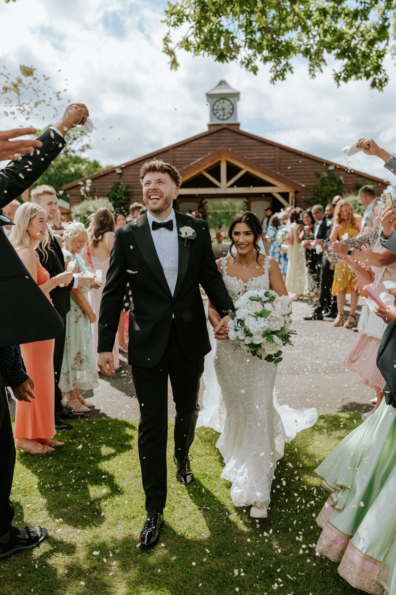 Joyous newlyweds walk through shower of confetti on garden path by rustic wooden chapel with clock tower.