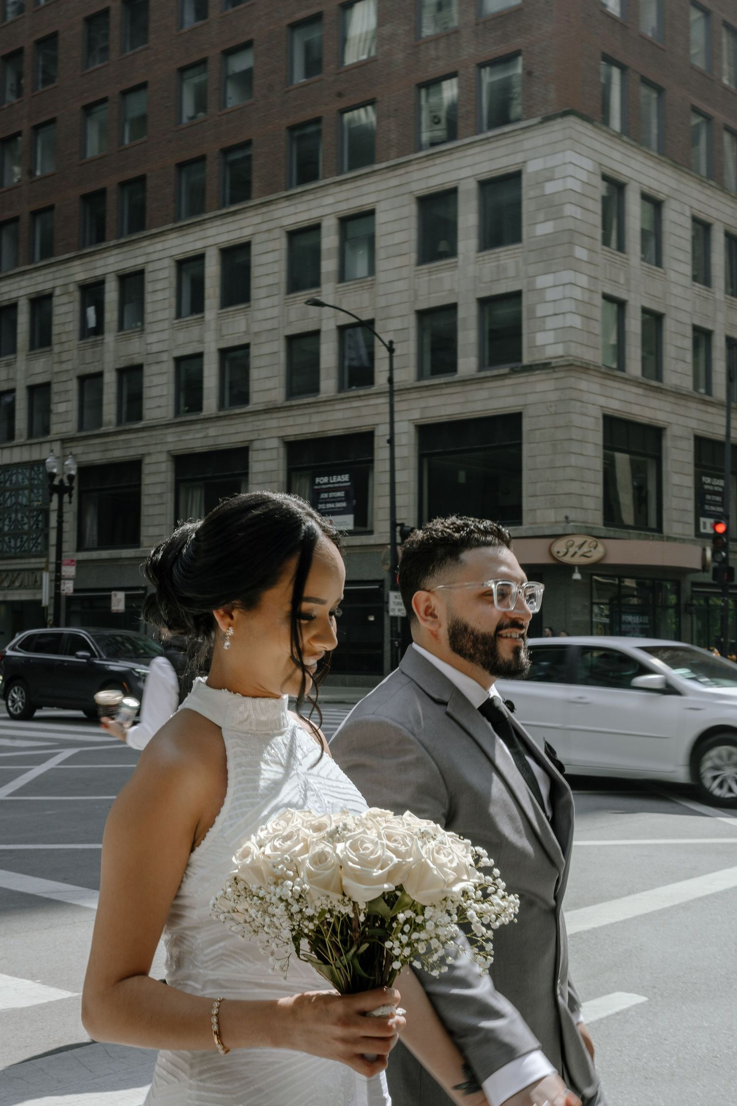 A couple walking through downtown city streets with a white bouquet against an urban brick building backdrop.