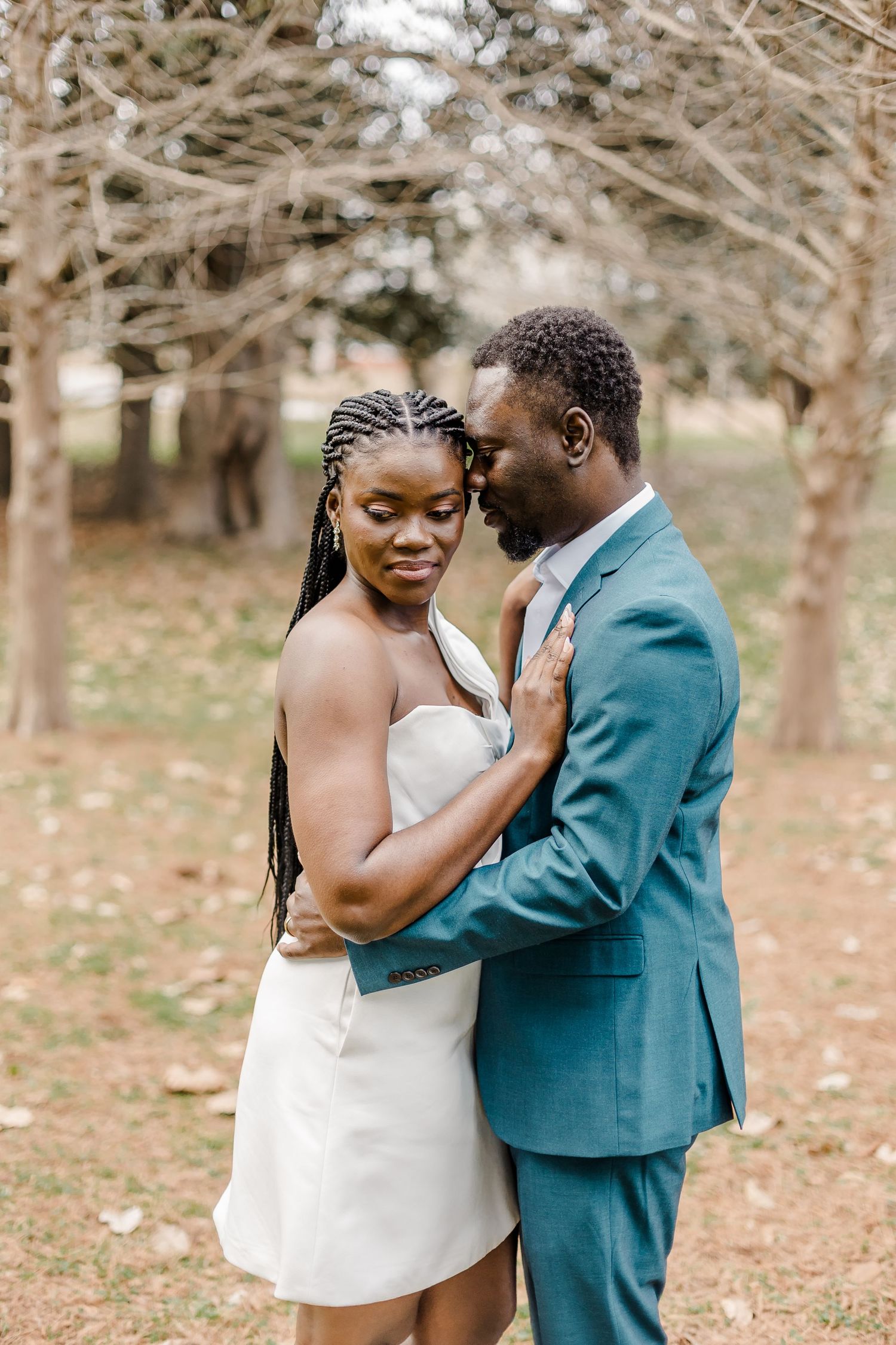 Couple in formal attire embraces in wooded setting during engagement or wedding shoot.
