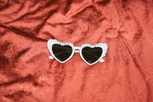 White heart-shaped sunglasses with black lenses placed on a red velvet textured background.